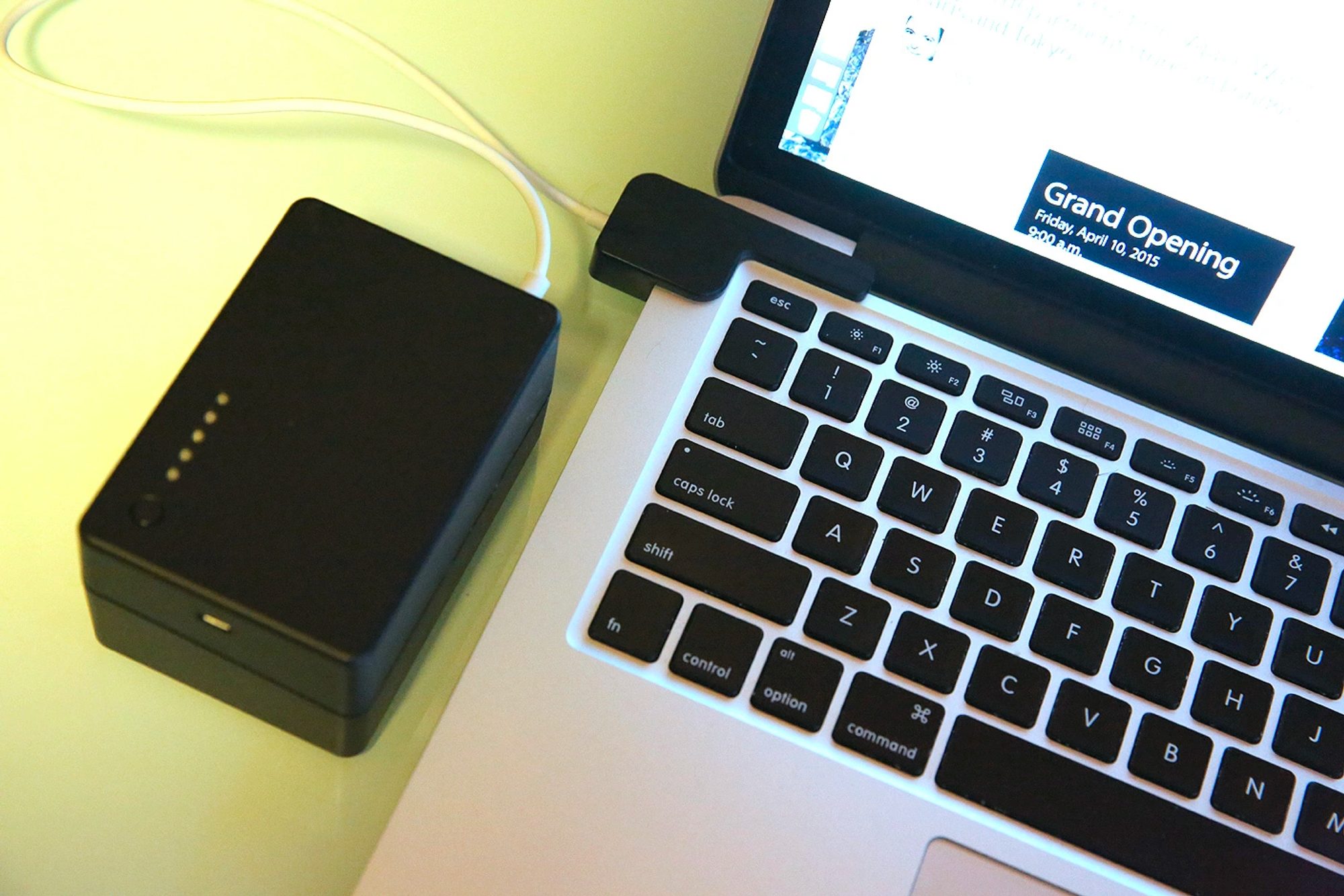 A black power bank with a row of charge indicator lights, cabled into a MacBook on a desk.