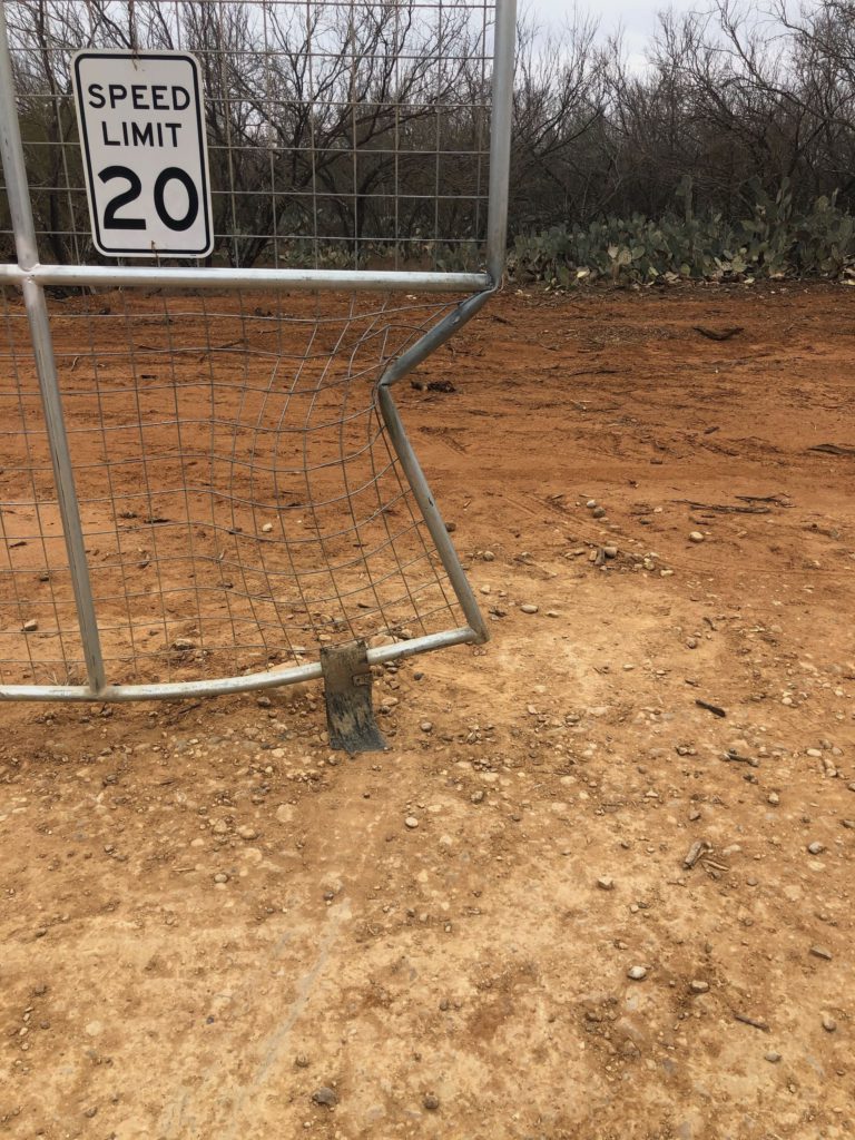 A wire gate propped in red dirt with a SPEED LIMIT 20 sign wired to it, prickly pear and bare brush behind.