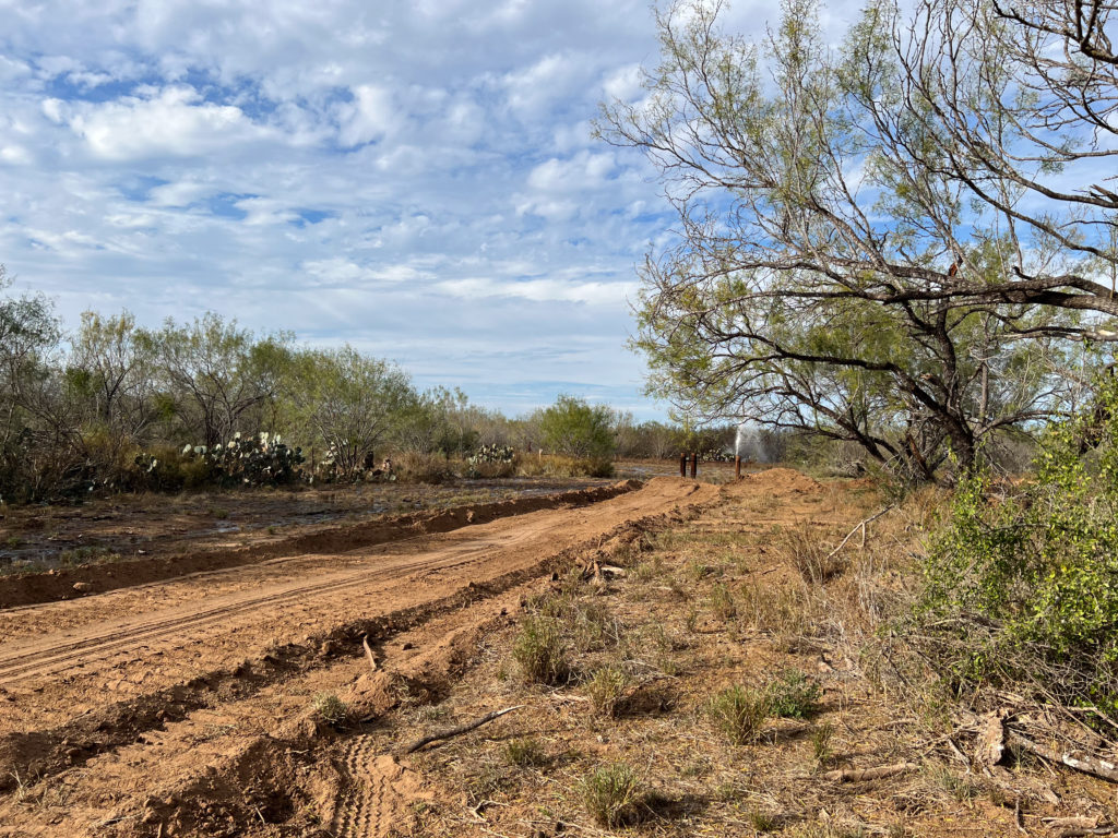 A freshly bladed red dirt road running alongside a cleared right of way, water spraying from a wellhead in the distance.