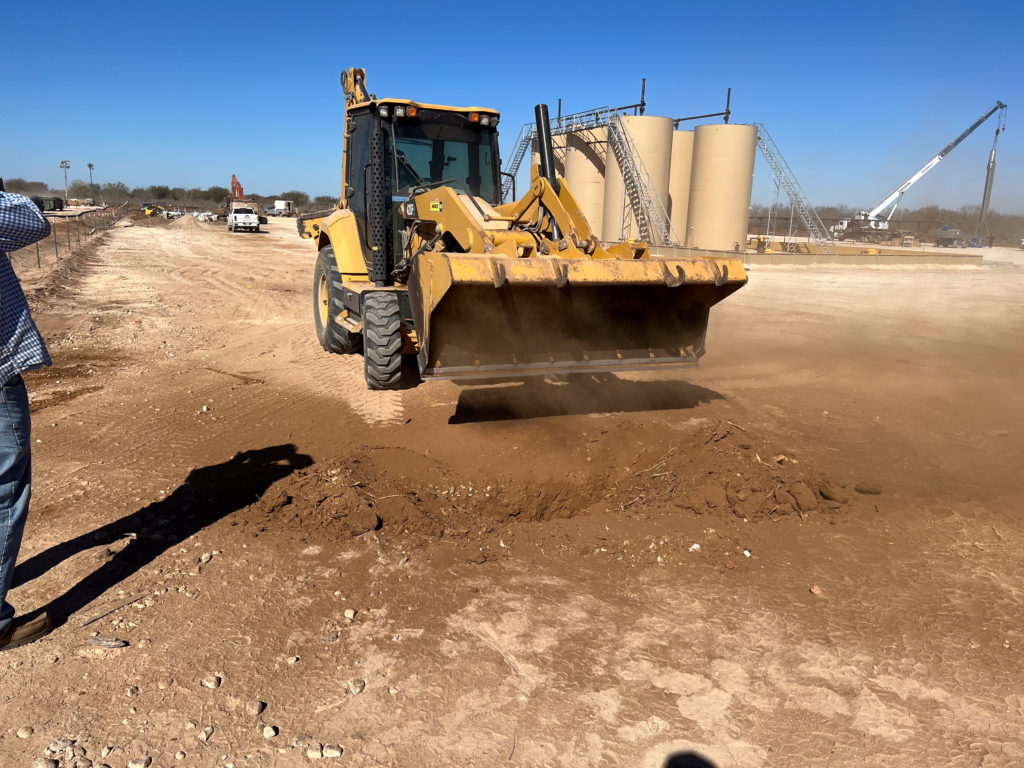 A backhoe grading a caliche pad in a cloud of dust, tank batteries and a crane working behind it.