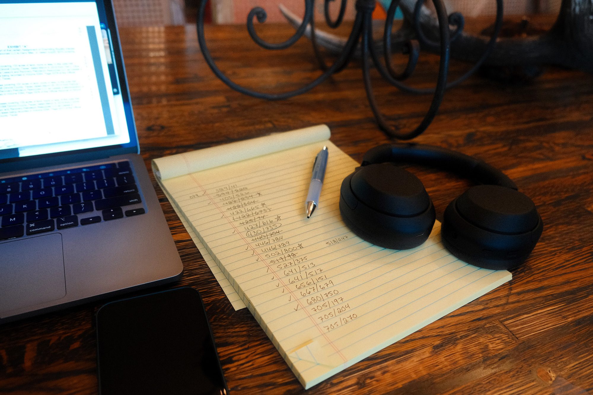 A laptop, a pen and headphones on a wooden table beside a yellow legal pad filled with handwritten volume and page numbers, many of them ticked off.