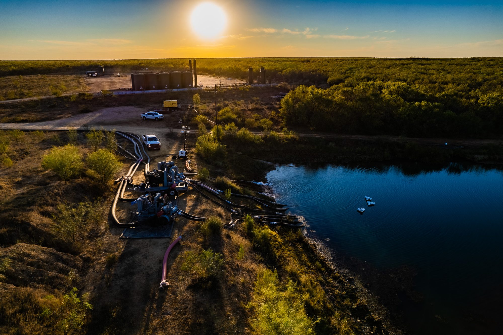 Aerial view at sunset of a water transfer setup, pumps on a pad with lay-flat hose running down to a pond and out toward a tank battery on the horizon.