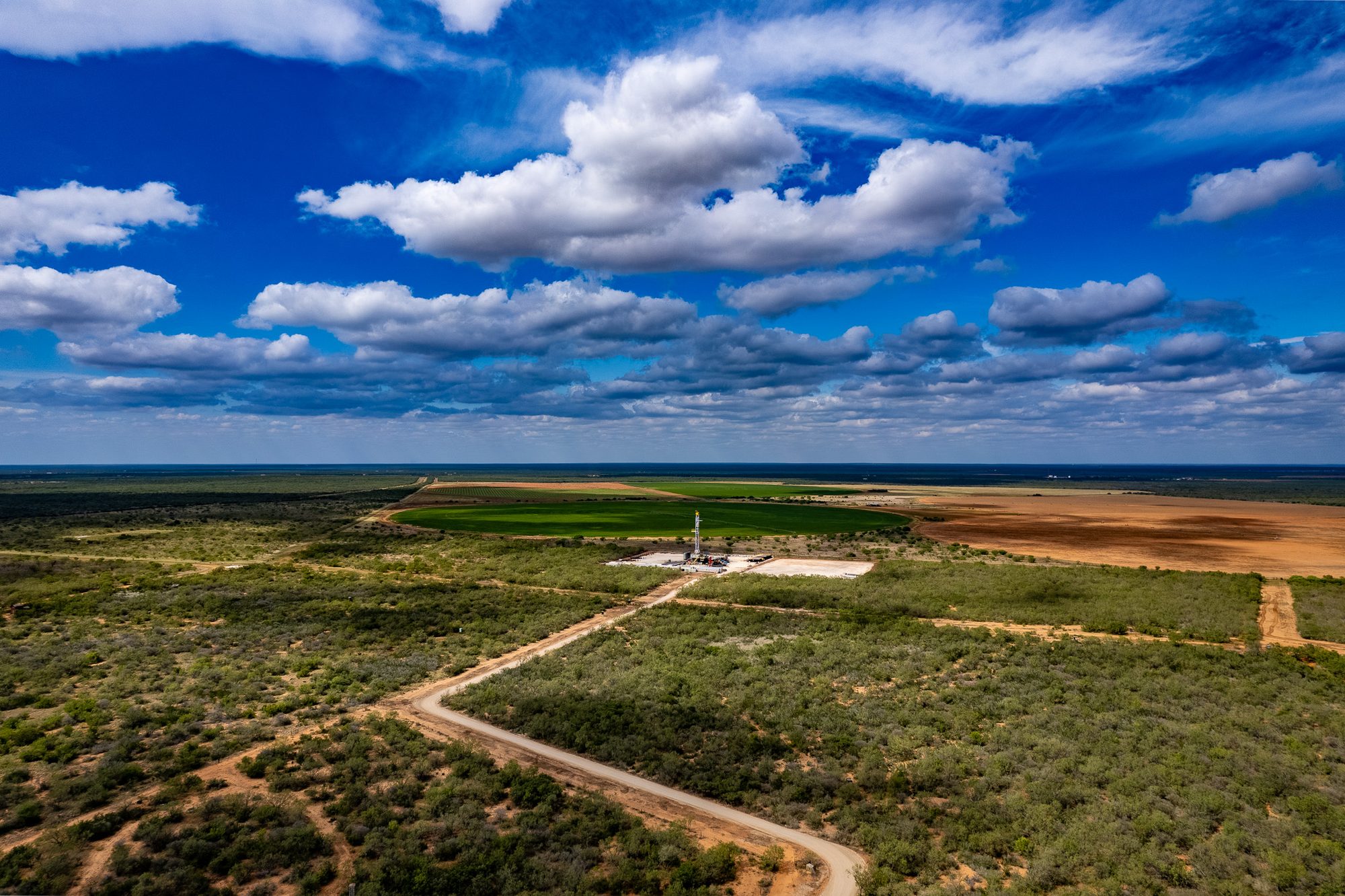 Aerial view over wide South Texas brush country, a small drilling rig on a pad in the middle distance and a green centre-pivot field beyond it.