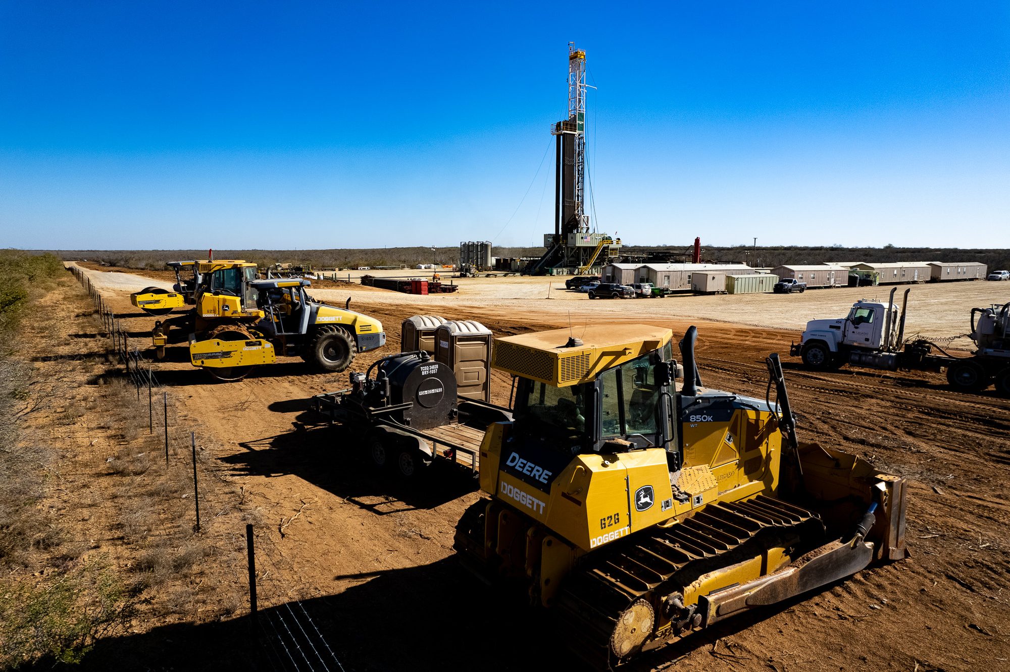 Aerial view of a drilling rig on a caliche pad with dozers, a compactor, portable toilets and a row of man camp trailers, brush country to the horizon.