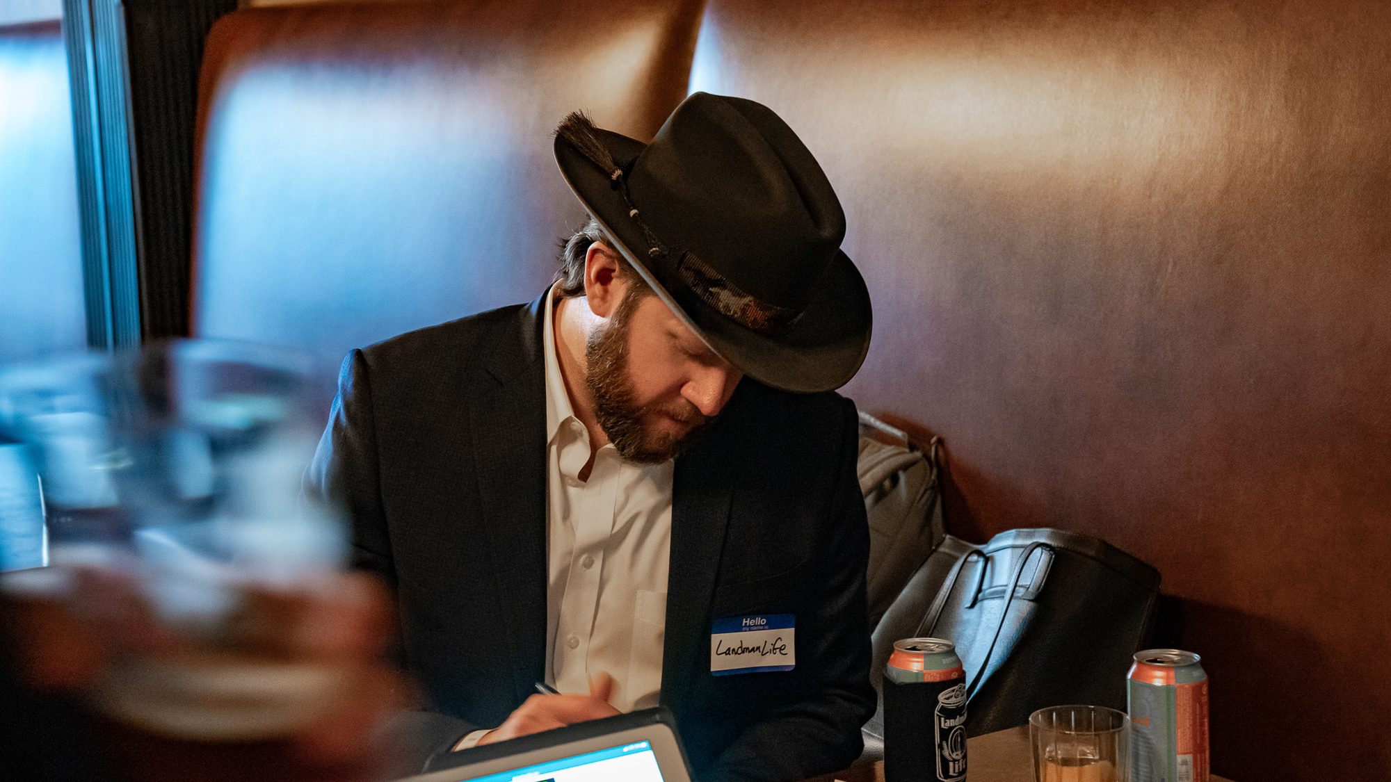 A man in a felt hat and blazer works on a laptop in a leather booth, wearing a Hello My Name Is nametag filled in as LandmanLife, beer cans in koozies on the table.