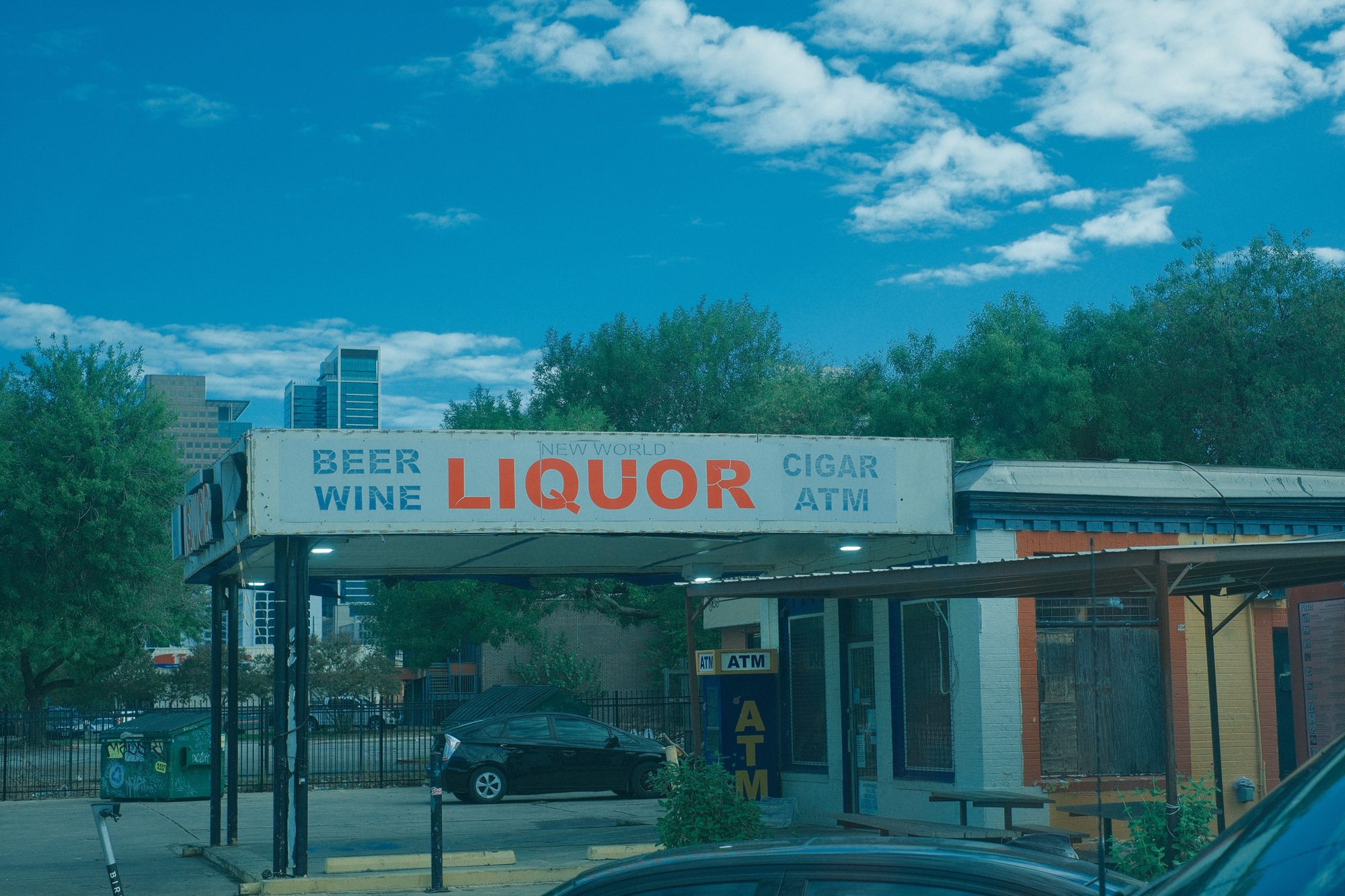 A New World Liquor storefront under a sign reading beer, wine, cigar and ATM, with a city skyline rising behind the trees.