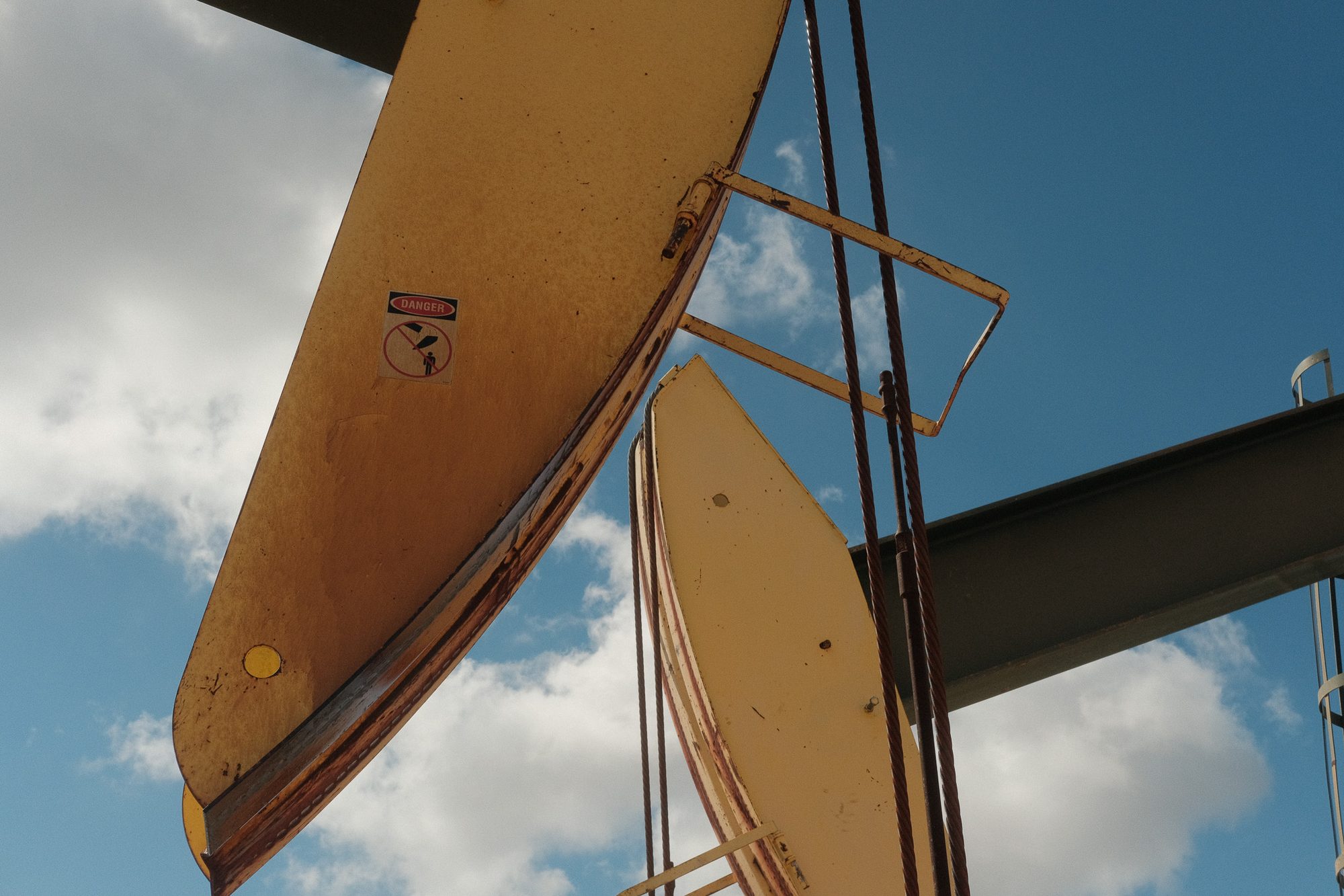Looking up at the counterweights of a pumpjack against a blue sky, a red DANGER sticker on the near weight.