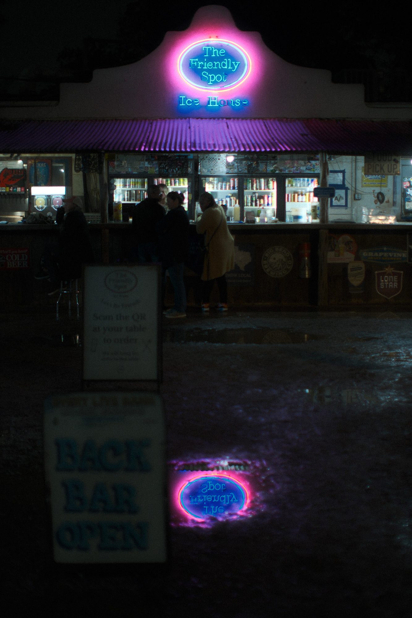 The Friendly Spot Ice House at night, its pink and blue neon sign reflected in a puddle in the foreground, people ordering at the lit bar behind.