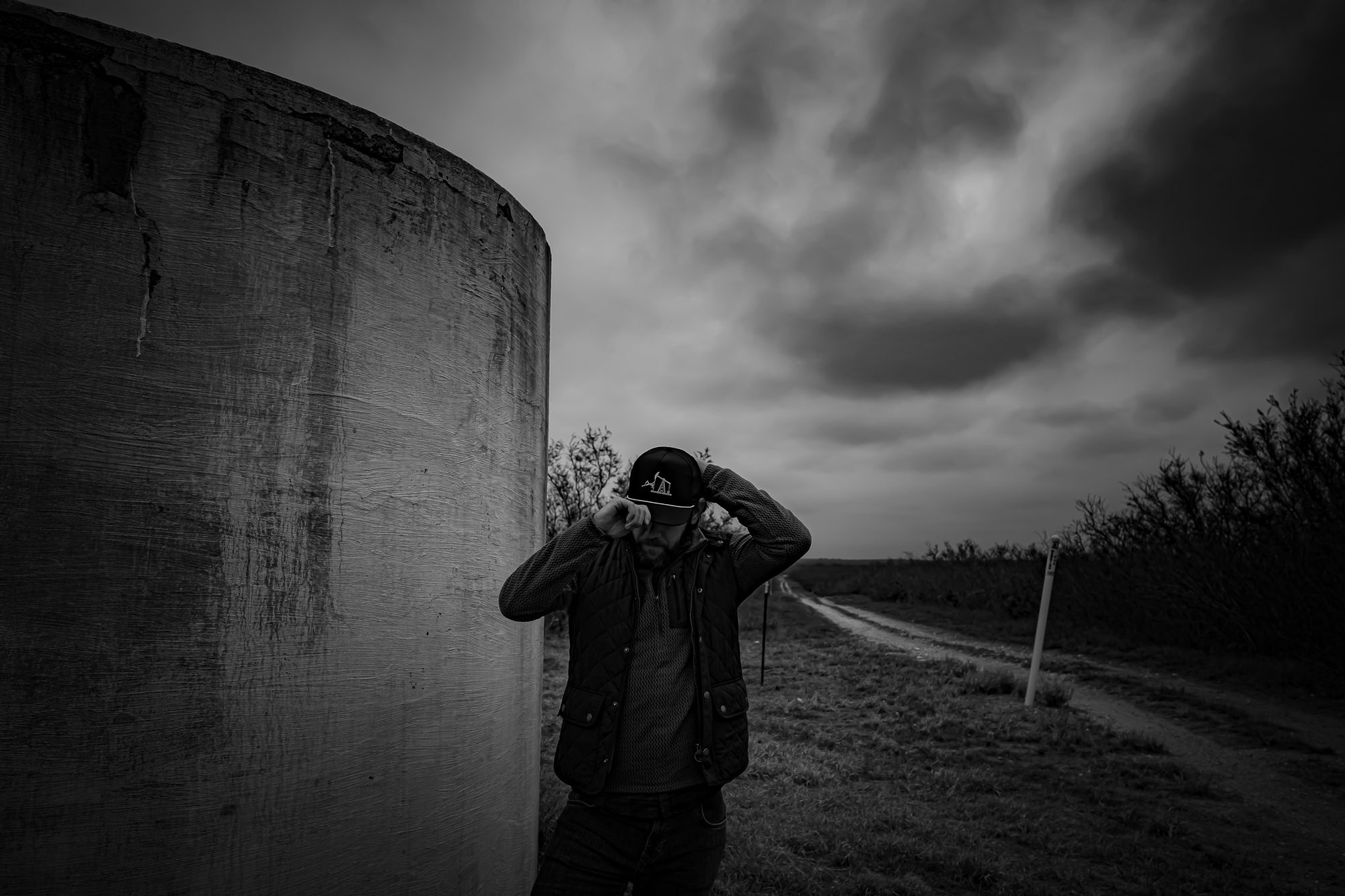 Black and white photograph of a man in a cap and quilted vest pulling his cap down, standing beside a concrete tank base under heavy storm cloud, a lease road running off behind him.