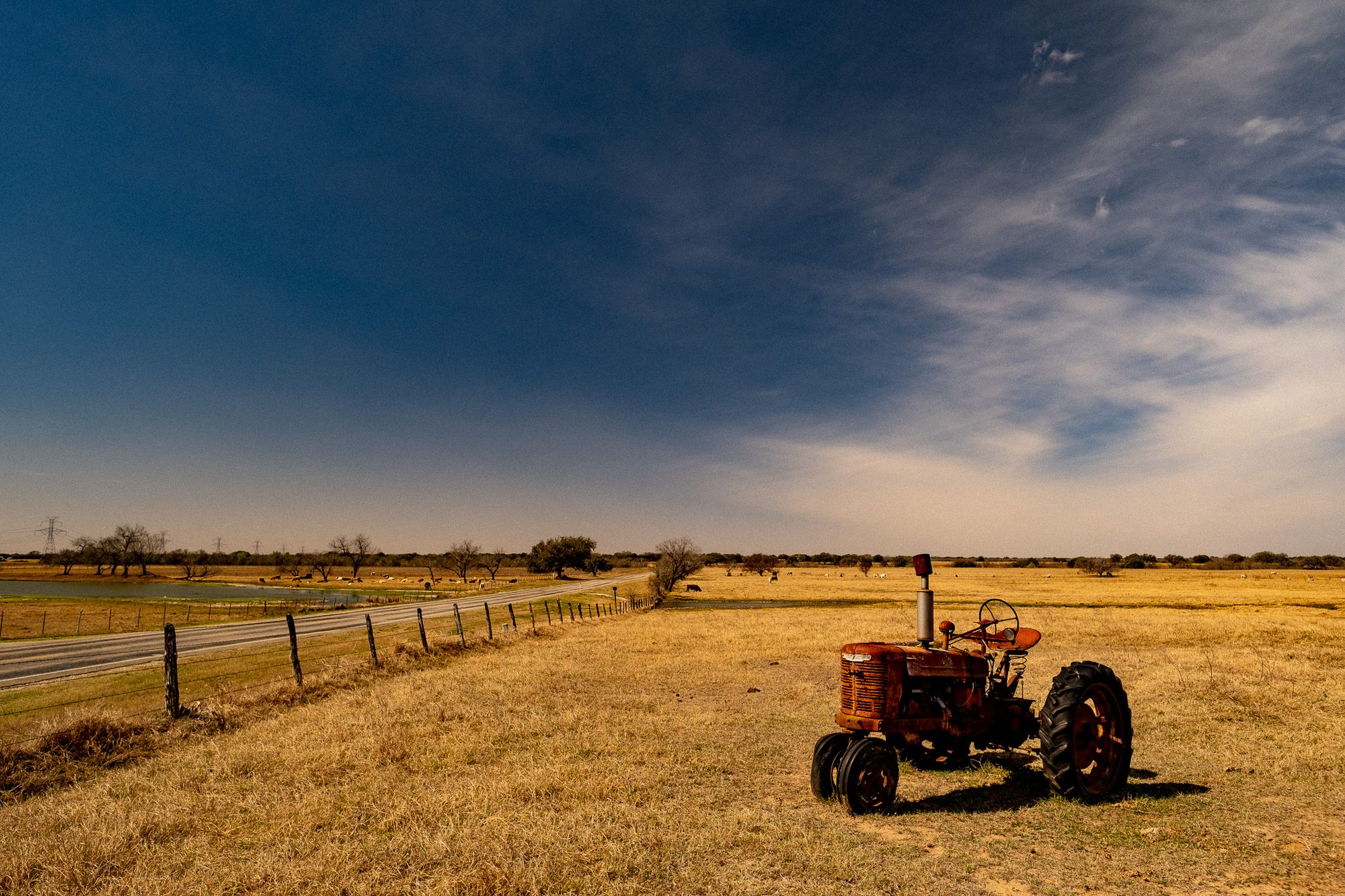 An old red tractor parked in dry pasture beside a two-lane road, cattle grazing near a stock tank in the distance.