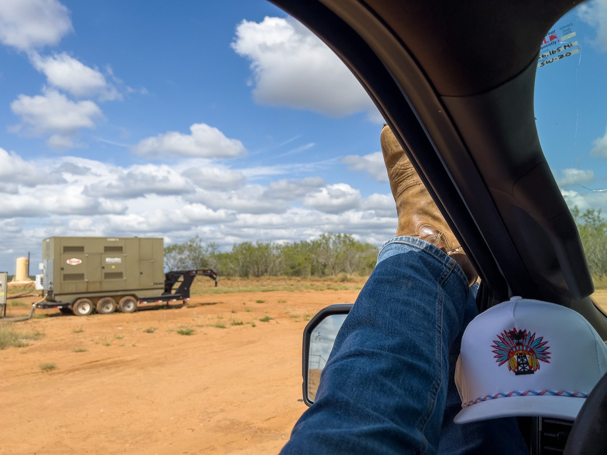 View from a truck cab with a boot propped on the door sill and a white cap with a headdress-and-derrick logo resting on the wheel, a trailer-mounted generator on the caliche pad outside.