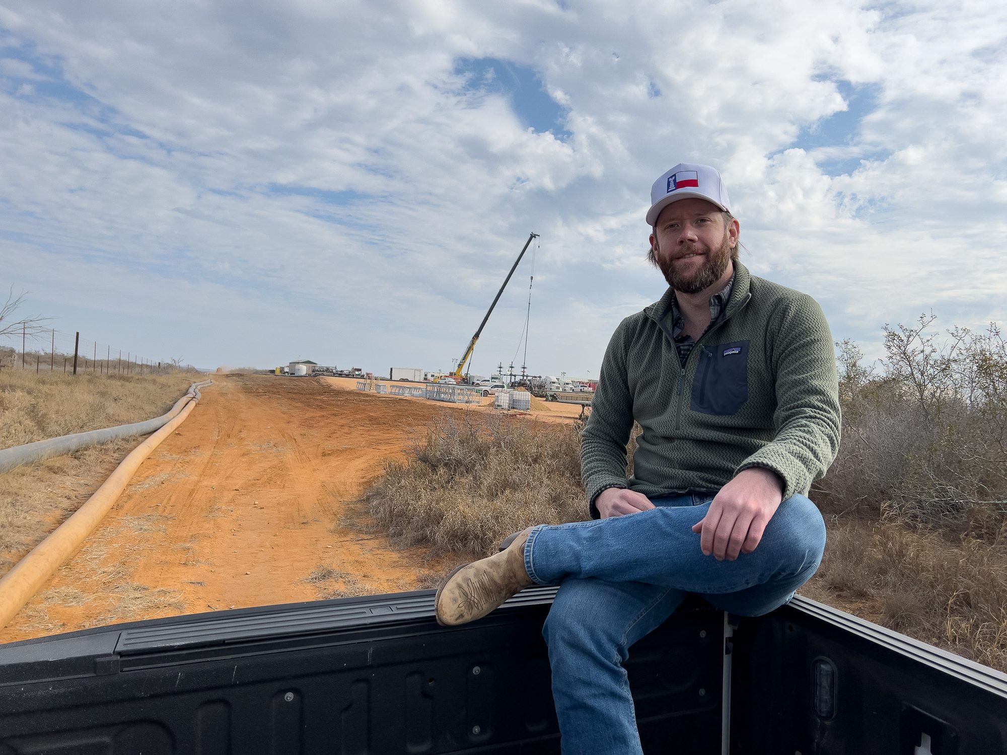 A man in a Texas flag cap and fleece sits on an open truck tailgate, a caliche road and a rig with a crane in the distance behind him.