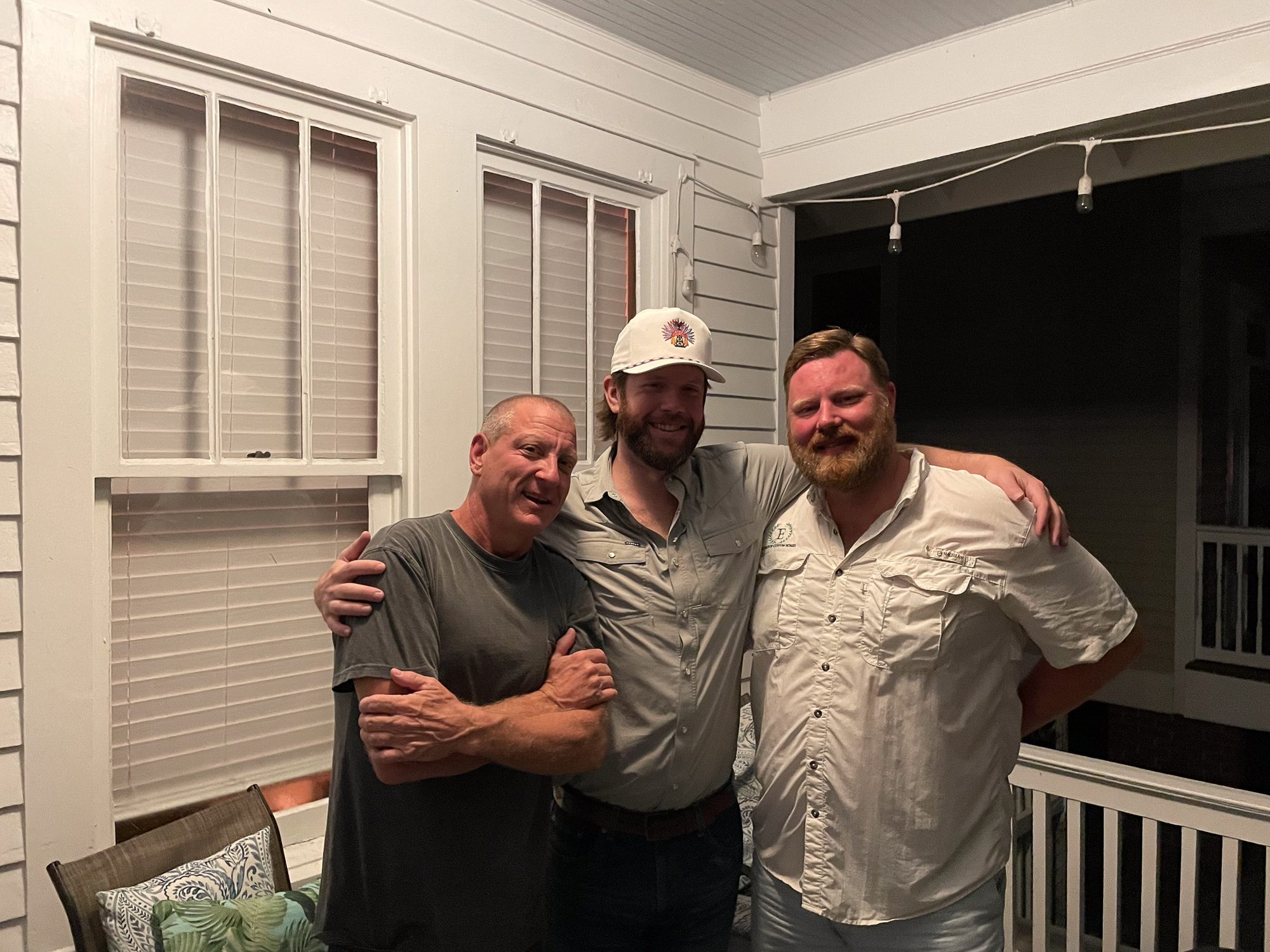 Three men stand arm in arm on a porch at night, all smiling at the camera.