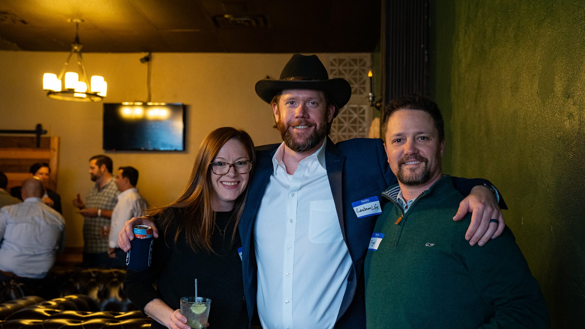 Three people pose together at an indoor event, drinks in hand and Hello My Name Is nametags on, one of them reading LandmanLife.