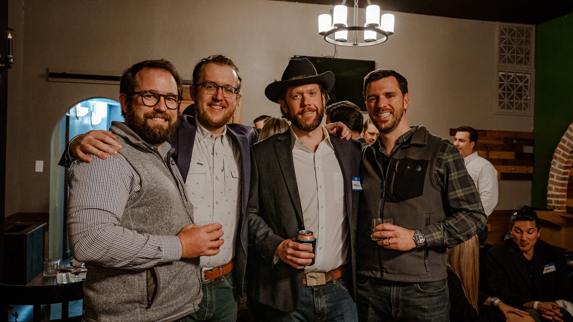 Four men pose together at an indoor event with drinks in hand, one wearing a felt hat and a LandmanLife nametag.