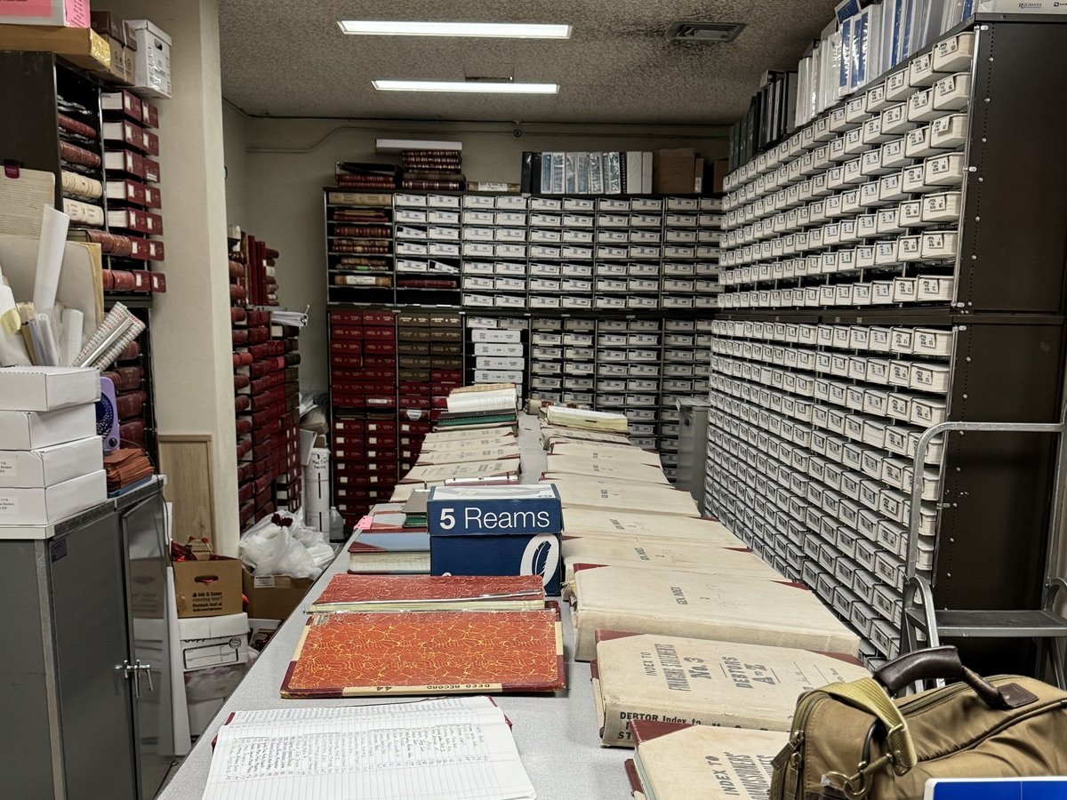 Shelves of bound record volumes lining a county records room, with books spread open on a work table
